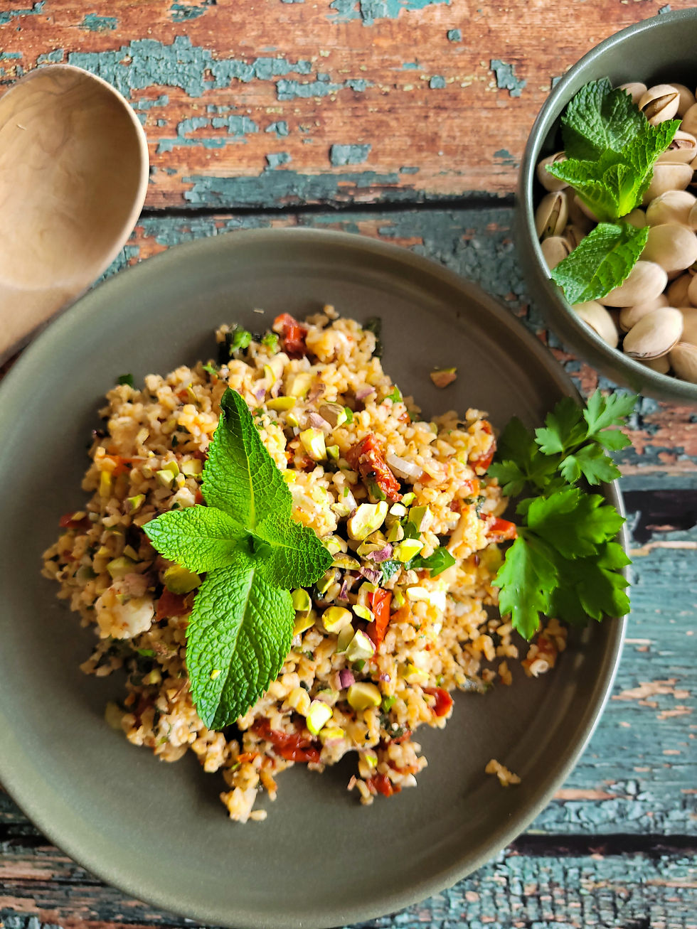 Bunter Bulgur-Salat mit Minzblättern und Petersilie auf grauem Teller, daneben Schale mit Pistazien. Rustikaler Holztisch im Hintergrund.