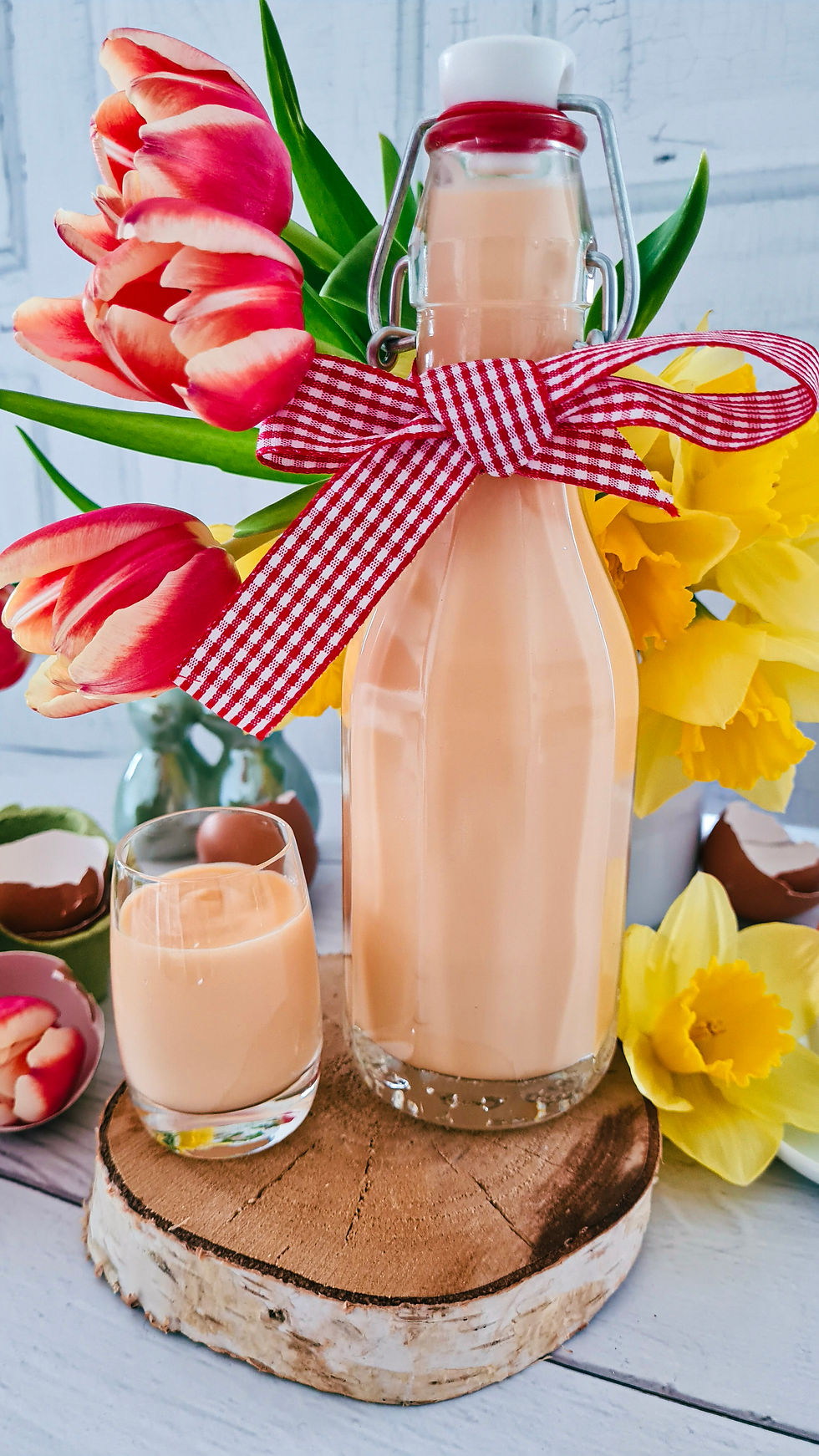 Flasche Eierlikör mit rot-weißem Band, Glas, rote Tulpen und gelbe Narzissen auf Holzscheibe vor hellem Hintergrund. Frühlingshaft.