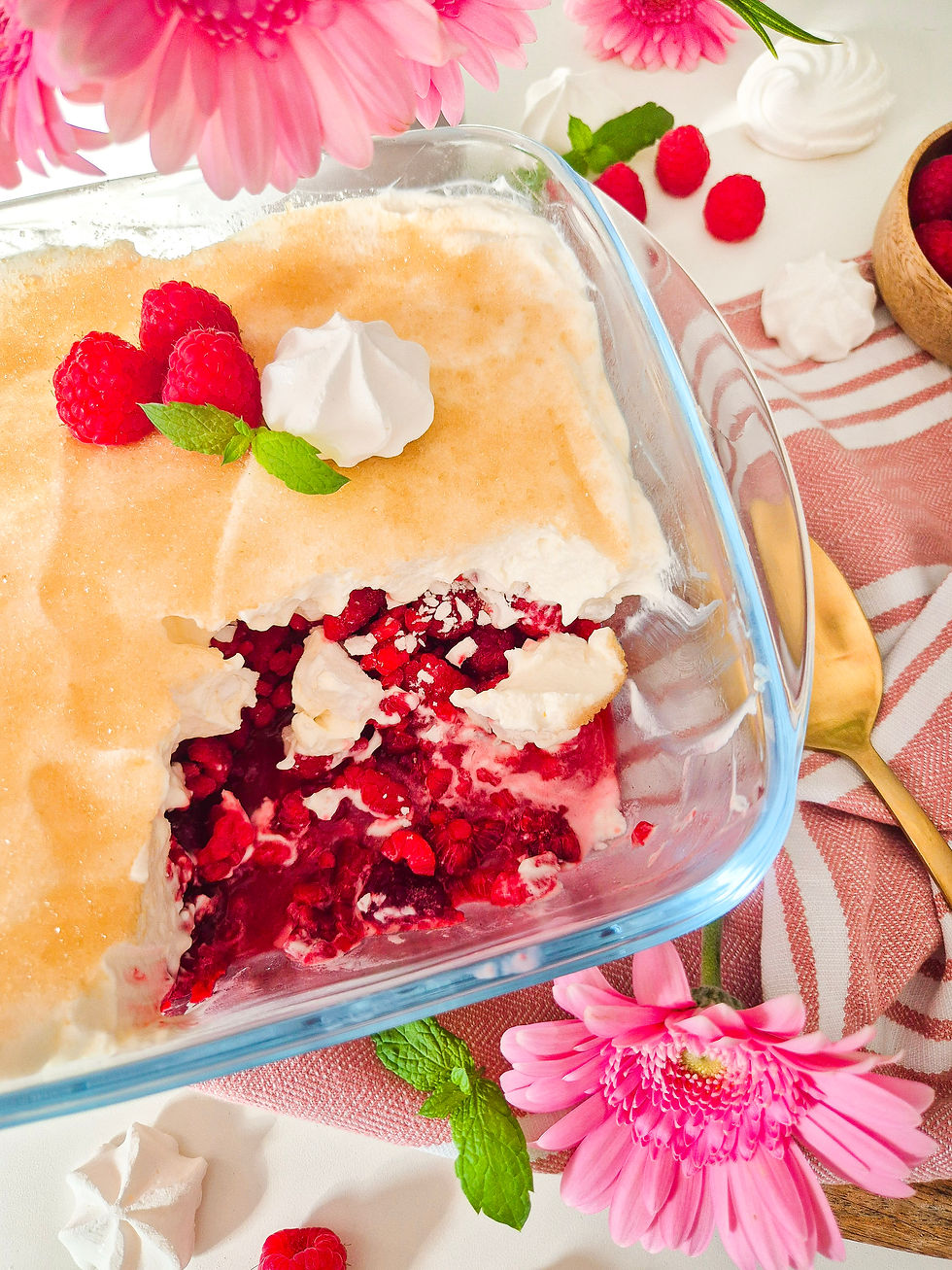 Glasform mit fruchtigem Himbeer-Dessert, Sahne, Baiser und Minze, umgeben von rosa Blüten auf weiß-rosa Tischdecke.