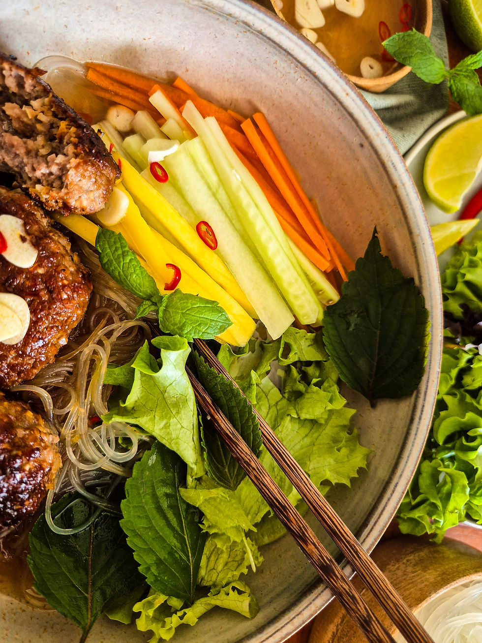 Bunte Nudel- und Fleischschale (Bun Cha) mit Gemüse, Kräutern, Limette und Essstäbchen auf rustikalem Tisch