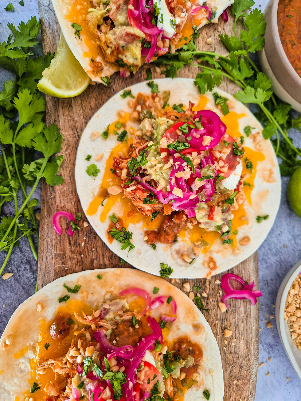 Drei Tortillas mit Pulled Chicken, Avocado, roten Zwiebeln und Koriander auf Holzbrett, mit Limettenspalte.