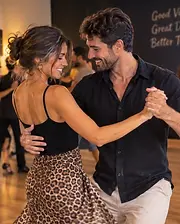 Smiling couple social dancing in a close partner hold, the woman in a leopard-print skirt, with other dancers in the background.