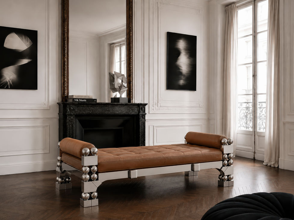 Orbital daybed in warm caramel leather and polished stainless steel, placed in a Parisian collector-style interior with classical boiserie, contemporary black and white photography, a black stone fireplace and mirror