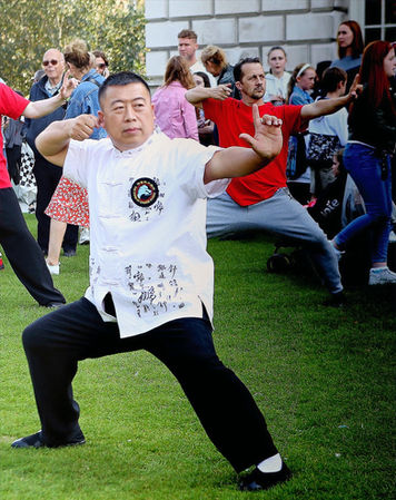 Grandmaster Zhu Chang Hai wearing white martial arts shirt and black trousers and his students in the background wearing red T-shirts are in a position called Shoot the arrow from 8 Piece Brocade (Baduanjin) and they are standing in a horse stance, right hand is pulling on the imaginary bowstring and left hand is extended to the side with the palm facing outwards and little finger, ring finger and middle finger are all bent and this was a part of Culture Night & Day event in front of the City Hall in Belfast, Northern Ireland.