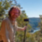 Smiling man with a camera on a stick in a scenic forest overlooking a river, under a clear blue sky. Brown shirt adds warmth to the scene.