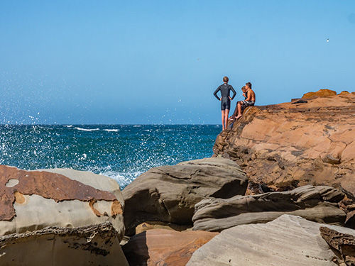 Three people on a rocky cliff by the sea, one standing with hands on hips, the other seated. Blue sky, waves splashing. Relaxed mood.