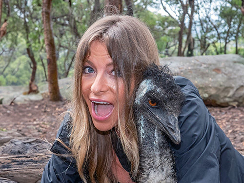 Woman hugging an emu in a forest setting, both appear surprised and joyful. Brown and green hues dominate with visible trees and rocks.
