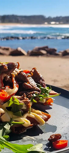 A plate of fresh salad with meat and sauce on a beachside table. Ocean waves, rocks, and a clear blue sky in the background. Vibrant and appetizing.
