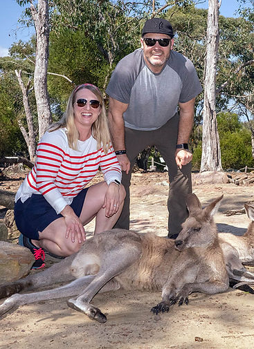 A couple in sunglasses pose with a resting kangaroo. Bushland background.