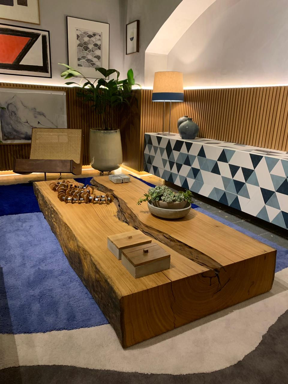 Mesa de centro em bloco maciço de madeira com objetos decorativos em uma sala com carpete azul e papel de parede em mosaico de triângulos