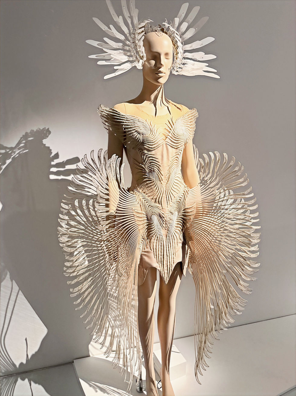 Mannequin in a beige avant-garde dress with feather-like sculpted wings, lit by sunlight against a gray wall.