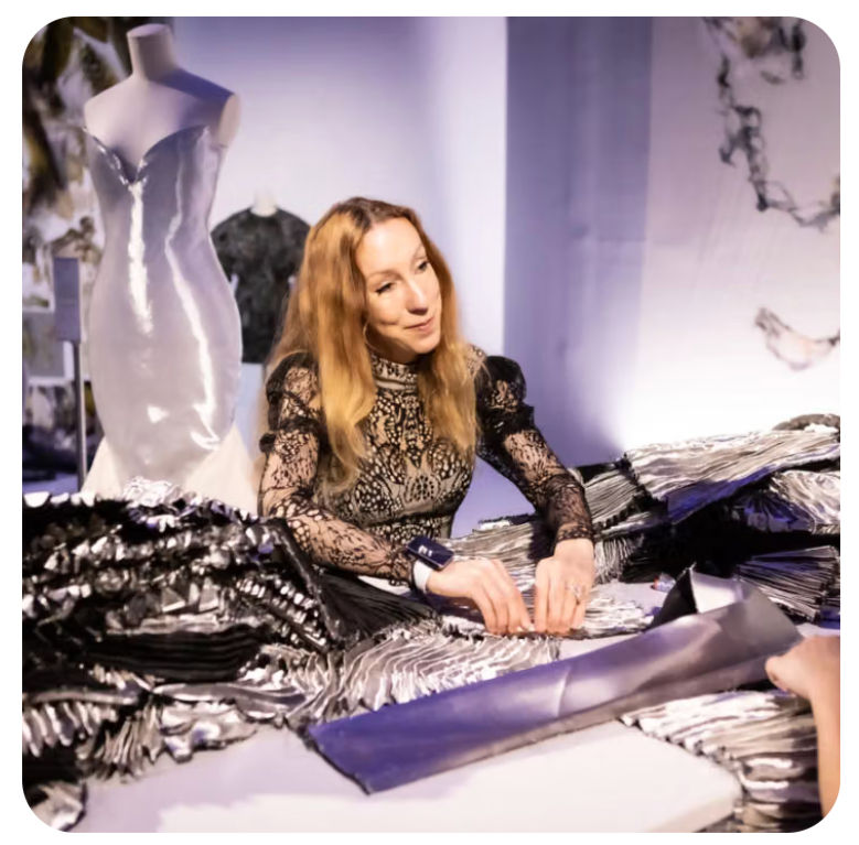 Woman in black lace dress works at a table of metallic fabric, with a mannequin dress behind her in a softly lit studio.