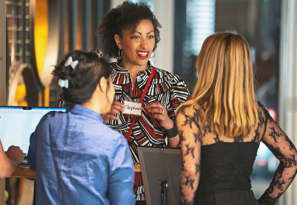 Three women in conversation indoors, one smiling and holding a name tag. Bright patterned shirt, laptop on desk, warm lighting.
