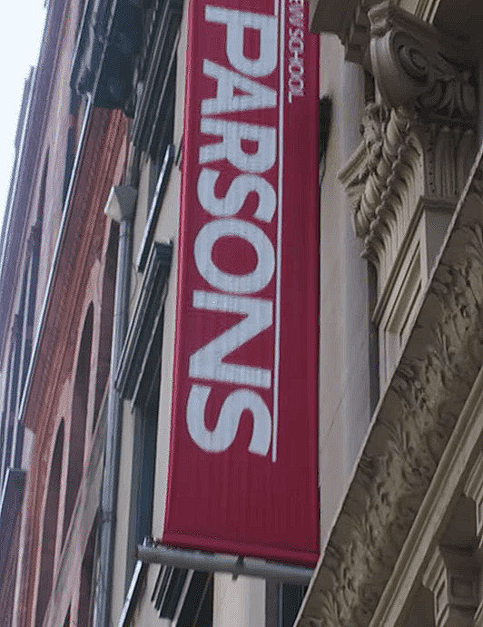Red vertical banner with "PARSONS" in white, hanging on a building with ornate details and brickwork. Urban street setting.