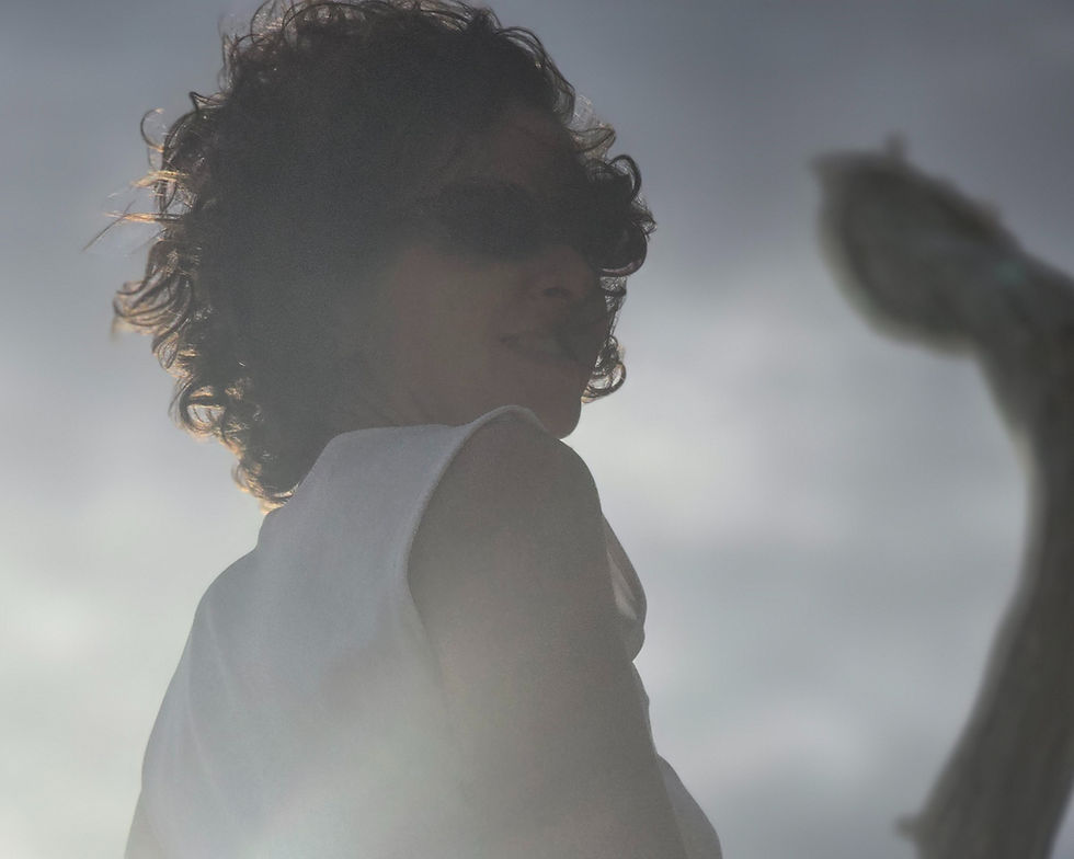 Woman with curly hair wearing sunglasses and a white top standing in sunlight, captured against a bright sky with soft lens flare.