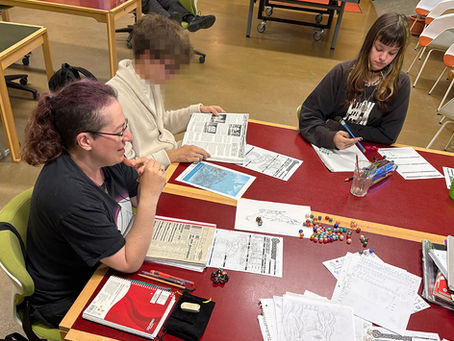 Pen & Paper bei Schule & Beruf Berlin: Teilnehmende sitzen mit einer Bildungsbegleitenden konzentriert an einem Tisch mit Charakterbögen, bunten Würfeln und Regelwerken bei einer gemeinsamen Rollenspiel-Runde.