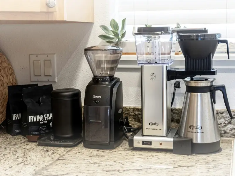 Coffee setup on a kitchen countertop with a grinder, scale, coffee bags, and a Moccamaster machine. Bright kitchen with a plant nearby.