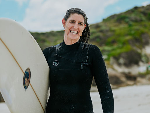 “Smiling woman in a wetsuit holding a surfboard, symbolising courage and starting before feeling ready.”