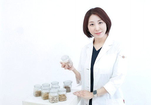 白衣の女性が室内で微笑む写真。手前に漢方の瓶が並ぶ背景画像