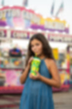 Girl in a blue dress sips lemonade from a yellow cup at a carnival booth, with a blurred Cotton Candy sign behind her.