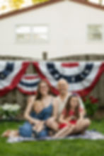 Smiling family of four sits on a blanket in a backyard, with red-white-blue bunting on a fence behind them.