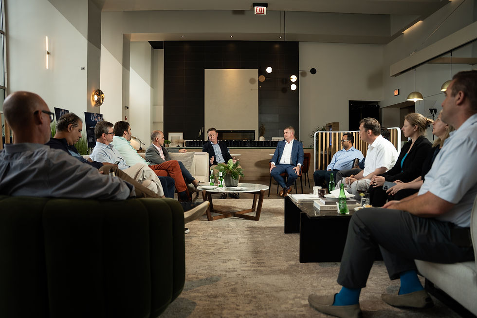 Business meeting in a modern lounge, with men and women seated in a circle listening to a speaker; EXIT signs visible.