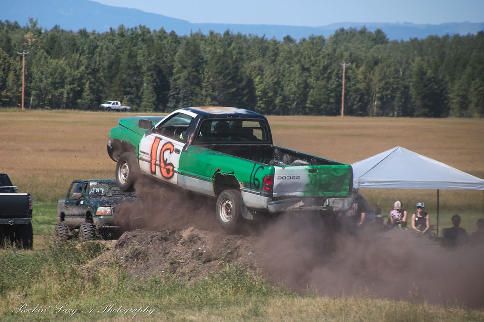 Green and white Dodge truck marked "16" jumps over dirt mound, kicking up dust. Spectators watch under a white tent in a grassy field.