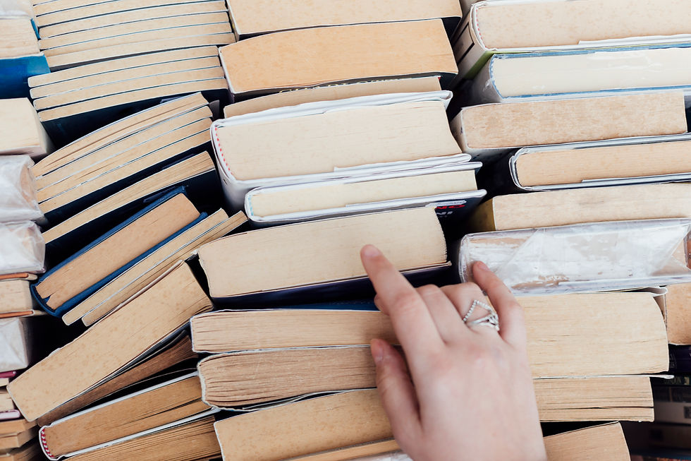 A hand with a ring reaches for a book among stacked, worn paperbacks. 