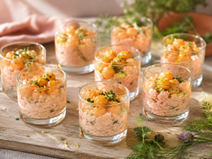Huit verrines apéritives en verre transparent remplies d'une préparation crémeuse rose pâle au crabe, garnies de billes de melon orange, d'herbes fraîches ciselées et d'un filet d'huile d'olive. Les verrines sont disposées sur une planche en bois clair, entourées de zestes de citron et d'herbes aromatiques, dans une lumière naturelle douce offrant une ambiance estivale, chaleureuse et élégante.