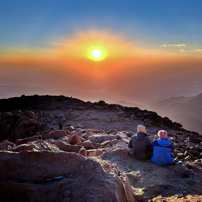 A couple sits on rocks at the top of Pikes Peak watching the sun rise over the clouds.