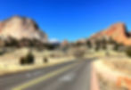 Road going into Garden of the Gods with large red rocks in the mid ground and Pikes Peak in the background on a sunny day.