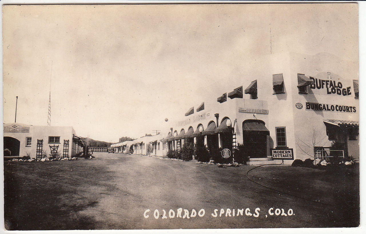 Historic photo of the Buffalo Lodge in Colorado Springs in black and white showing the main lodge on the right and empty street going to the rooms with Colorado Springs, Colo in white in the bottom center of the image.