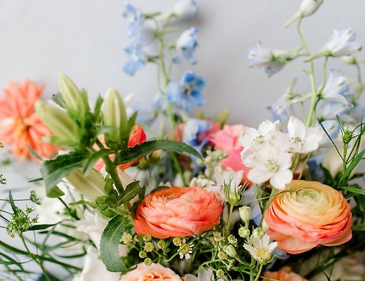 Close-up of a wedding bouquet featuring peach ranunculus, white scabiosa, pale blue delphinium, and fresh greenery in a loose