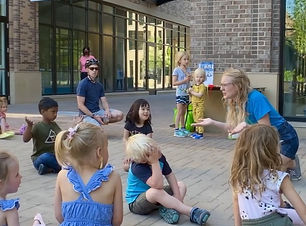 Madeline kneels in front of a group of young people at an outdoor plaza. She has her arms out to the side and is making a questioning face. One child mimics this and another child laughs.