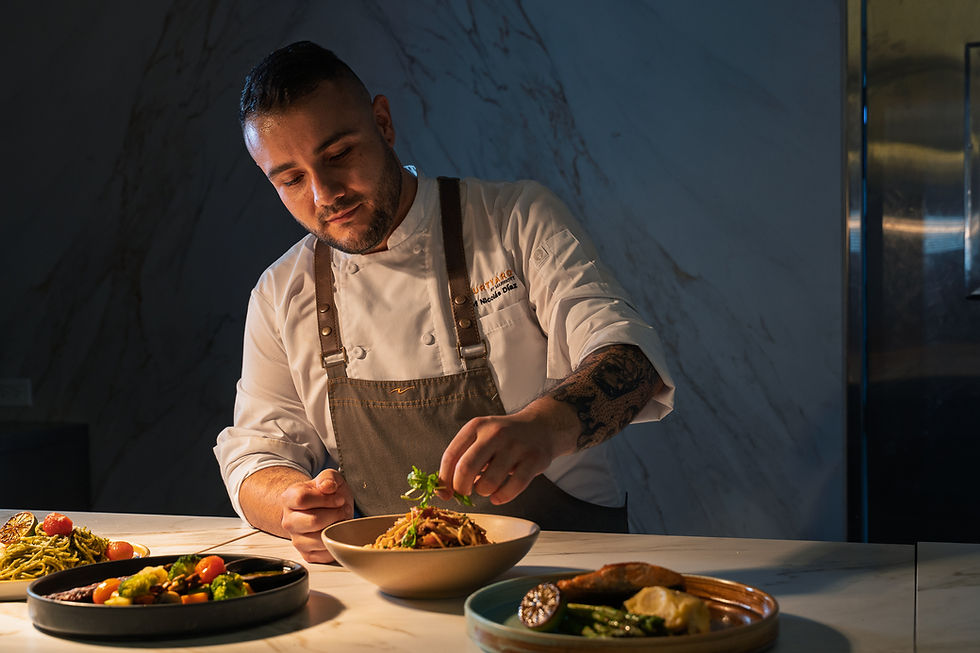 Chef en cocina elegante, decorando un platillo. Lleva uniforme blanco y delantal marrón. Varios platos coloridos en la mesa de mármol.