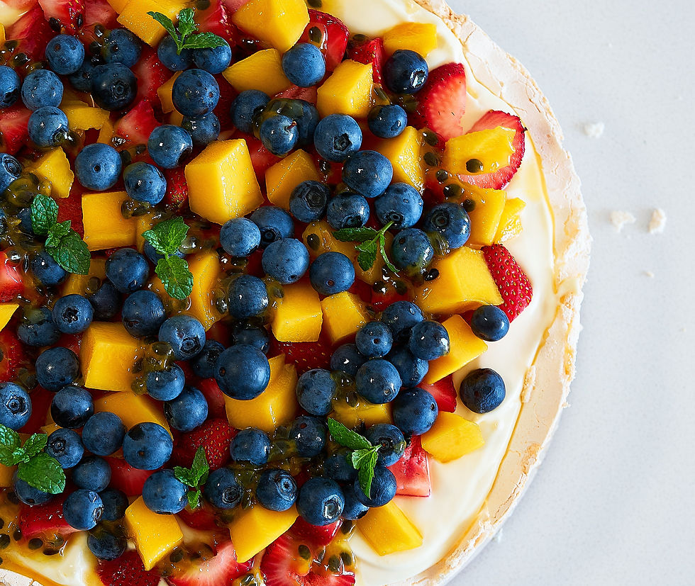 Pavlova decorada con arándanos, mango, fresas y menta sobre crema blanca. Frutas vibrantes en un fondo claro, transmite frescura y dulzura.