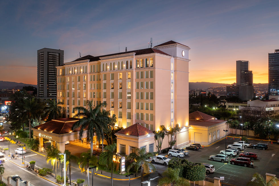 Hotel iluminado en ciudad al atardecer, con palmeras y coches estacionados. Cielo anaranjado y rascacielos en el fondo. Ambiente tranquilo.