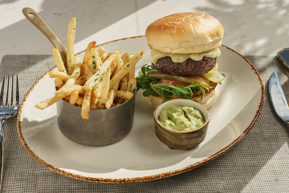 Hamburguesa con queso y vegetales en un plato junto a papas fritas en un recipiente metálico y guacamole. Fondo claro, ambiente casual.