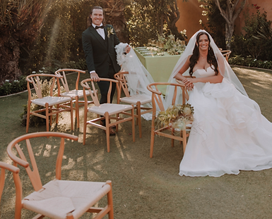 Close‑up of Wishbone elegant event chairs arranged around a table, each with neutral upholstery and refined wooden frames. The chairs are staged outdoors against a lush greenery backdrop