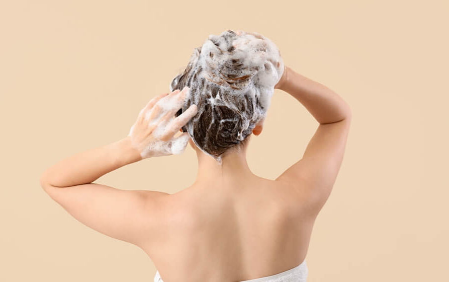 Person with a towel wrapped around body washes hair lathered with foam against a plain beige background. Mood is calm and refreshing.