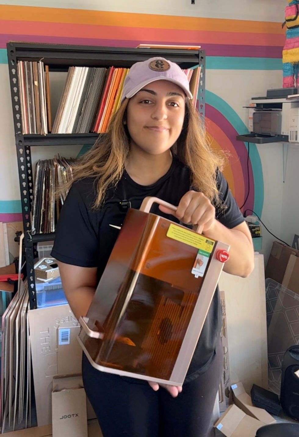 Jessica holding XTool F2 Laser while smiling, standing inside her she shed wearing a black shirt with a purple baseball cap with her logo engraved on it