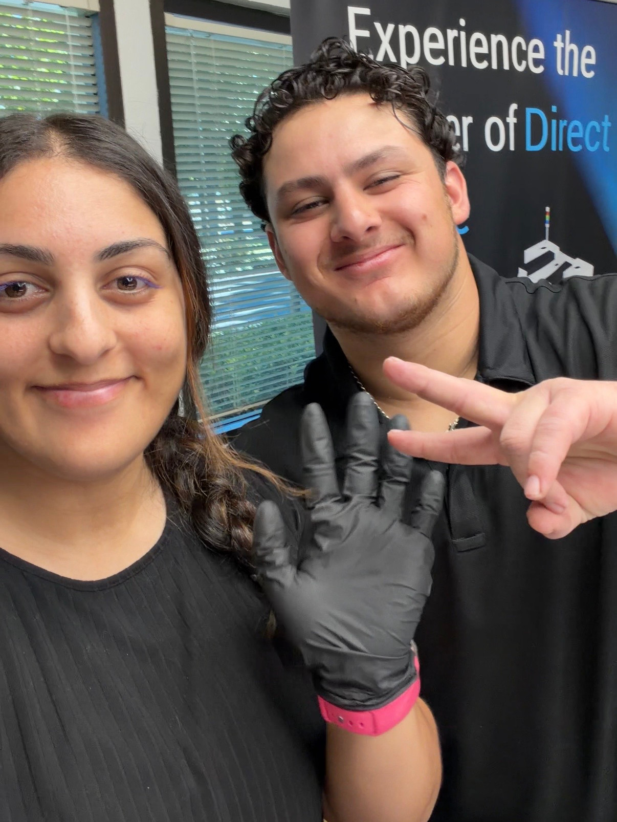 Smiling two young adults take a selfie indoors; one flashes a peace sign and wears a black glove.