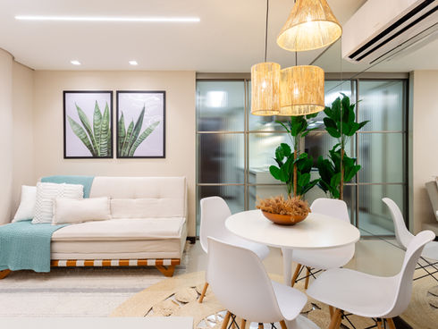 Interior do loft escandinavo com decoração minimalista e paleta de cores em tons de off white. À esquerda, um sofá bege com almofadas brancas e uma manta azul clara. Ao centro, uma mesa redonda branca cercada por cadeiras brancas modernas. Acima, três luminárias pendentes com cúpulas de fibras naturais iluminam o espaço. À direita, uma grande planta verde em vaso adiciona um toque de frescor tropical. No fundo, divisórias de vidro canelado separam sutilmente os ambientes. O chão é coberto por um tapete claro com detalhes geométricos e uma textura natural.