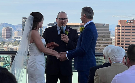 A bride in a white dress and veil and a groom in a blue suit hold hands during an outdoor wedding ceremony. An officiant stands between them, speaking into a microphone. The backdrop features city buildings, conveying a joyful and formal atmosphere.