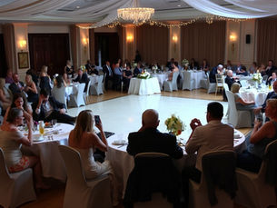 A wedding reception with guests sitting at round tables around an empty dance floor. The guests look bored, confused, and disengaged. Some are staring at their phones, others are chatting quietly without much energy. The lighting is dim, and the mood is dull. The room is decorated for a wedding with white tablecloths, floral centerpieces, and string lights, but the overall vibe feels awkward and lifeless.