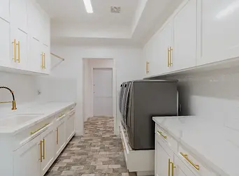 A remodeled laundry room with white walls, counters, and cabinets, a sink, gold hardware, and gray appliances