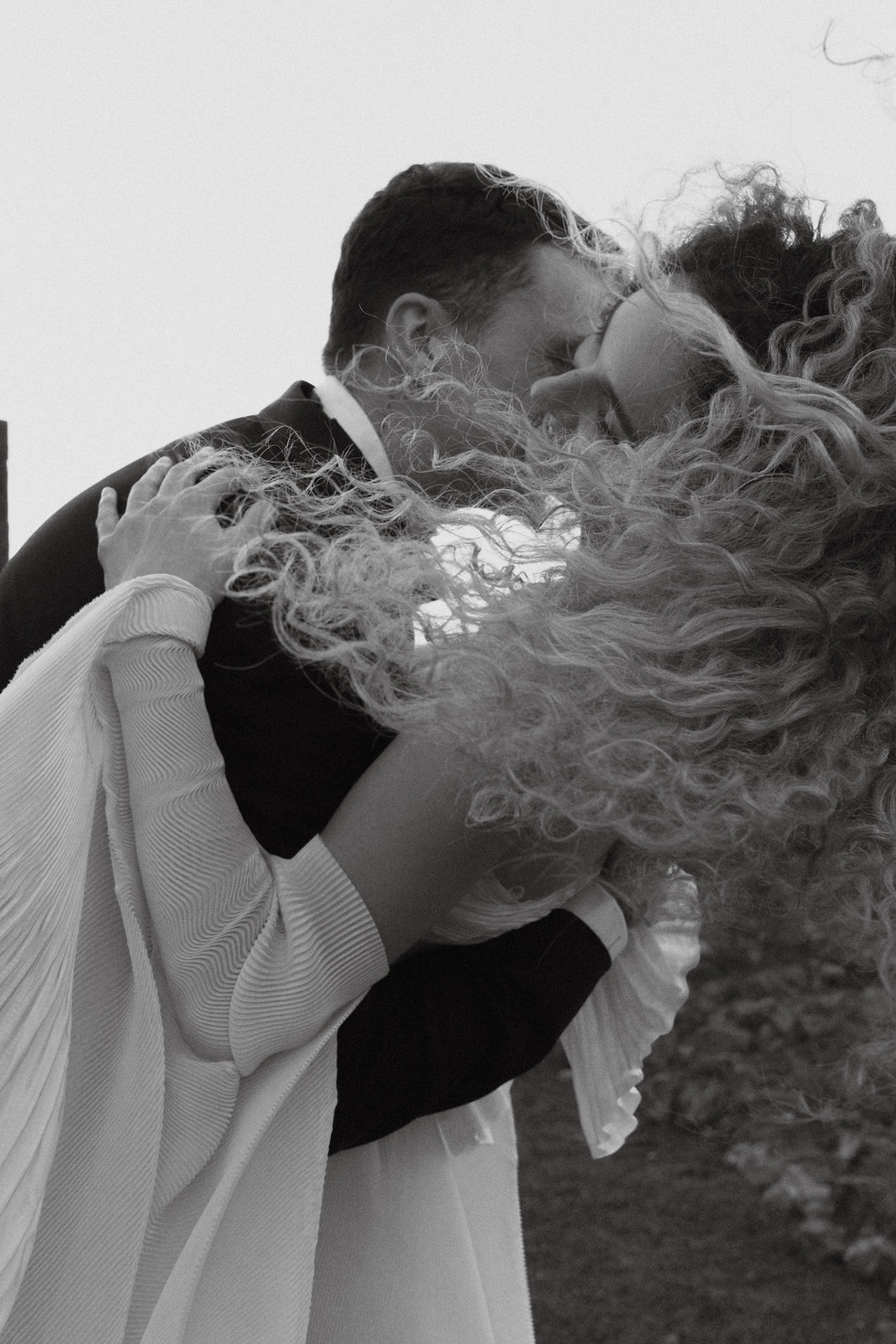 a black and white photo of the groom kissing the bride while bride's hair is flowing with the wind