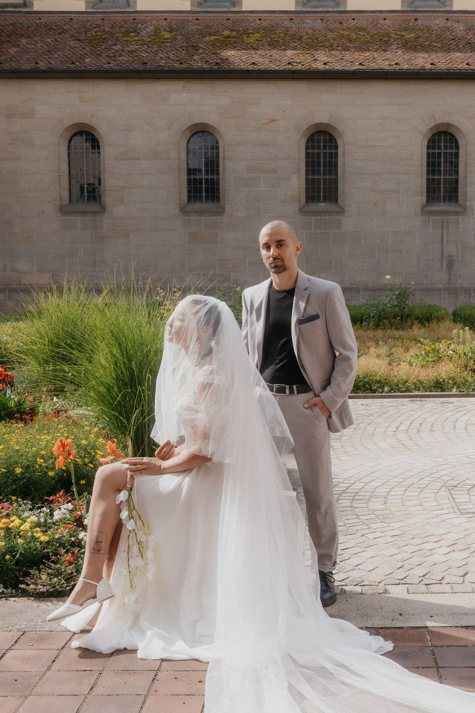  bride sitting on a stone in front of the church with groom standing next to her in Heilsbronn