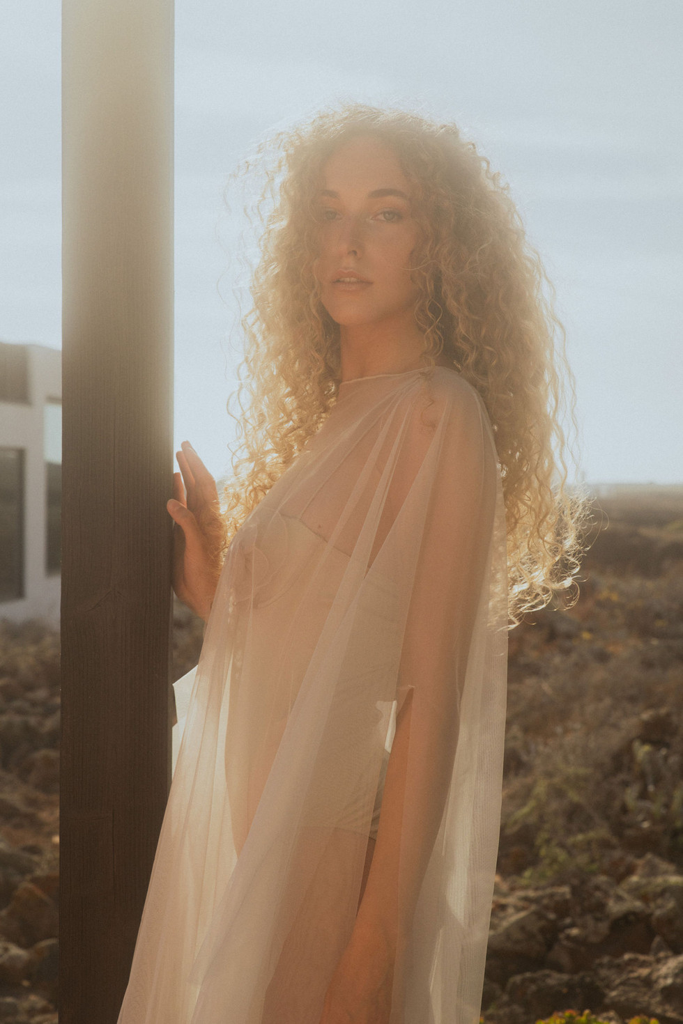Bride bathed in golden hour light, wearing a sheer, flowing gown with soft curls cascading over her shoulders.