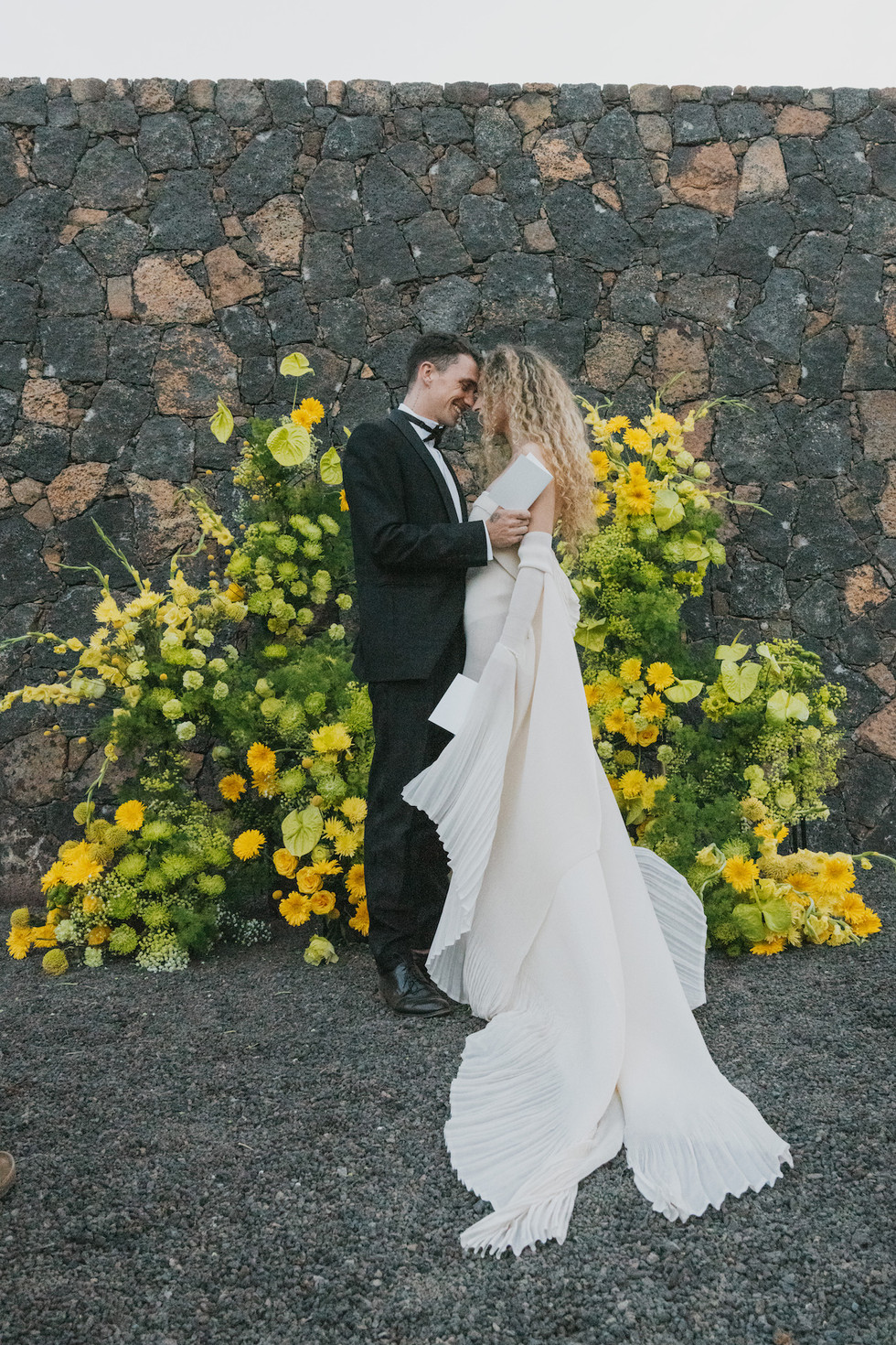 Groom reading vows as the bride leans in smiling, framed by vibrant yellow floral arrangements and textured walls.